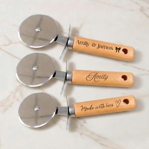 Könnte beinhalten: Drei Pizzaschneider aus Edelstahl mit hellen Holzgriffen. Jeder Griff ist mit einer Lasergravur versehen: oben steht Amily & James mit einer Schleife, in der Mitte Amily und unten steht Made with love mit einem Herzsymbol.