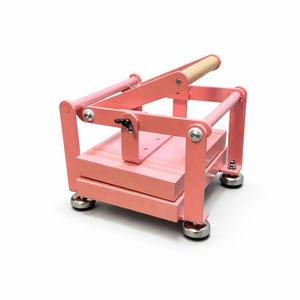 May include: A pink craft press with a wooden handle. The press has a rectangular base and a hinged top, supported by metal bars. The press is designed for crafting and has a sturdy construction.