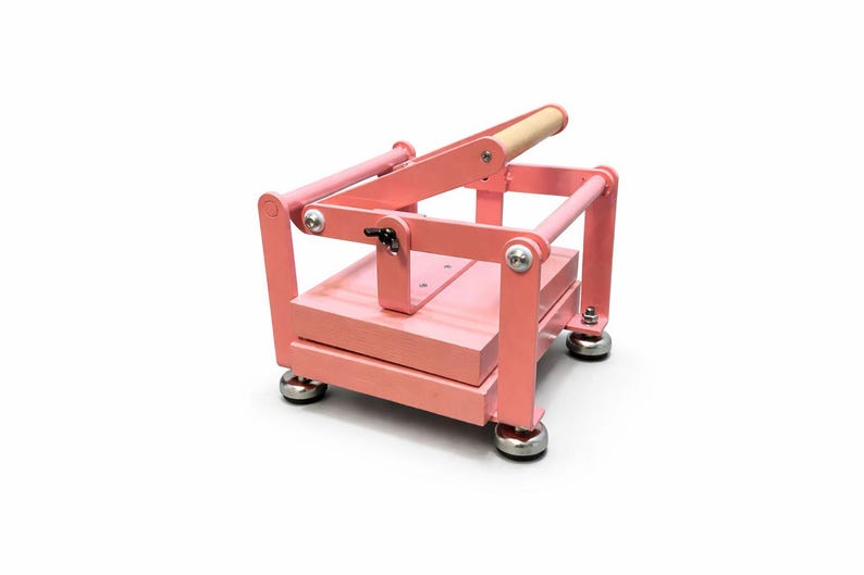 May include: A pink craft press with a wooden handle. The press has a rectangular base and a hinged top, supported by metal bars. The press is designed for crafting and has a sturdy construction.