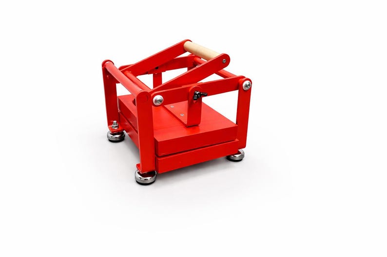 May include: A bright red wooden press with a natural wood handle. The press has a rectangular base and a hinged top, with silver hardware. The press is designed for a specific purpose, likely for crafts or food preparation.