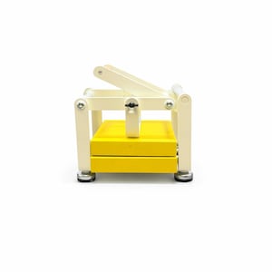 May include: A yellow and cream-colored wooden press with a hinged top. The press has a rectangular base and a lever arm. The base is yellow, and the frame is cream-colored. The press is designed for crafting or other similar activities.