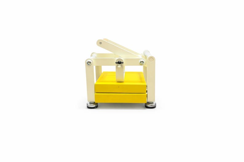 May include: A yellow and cream-colored wooden press with a hinged top. The press has a rectangular base and a lever arm. The base is yellow, and the frame is cream-colored. The press is designed for crafting or other similar activities.