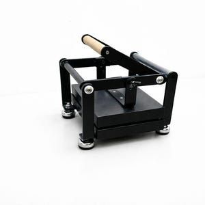 May include: A black book press with a wooden handle. The press has a rectangular base and a hinged top, with silver-colored hardware. The press is designed for crafting or bookbinding projects.