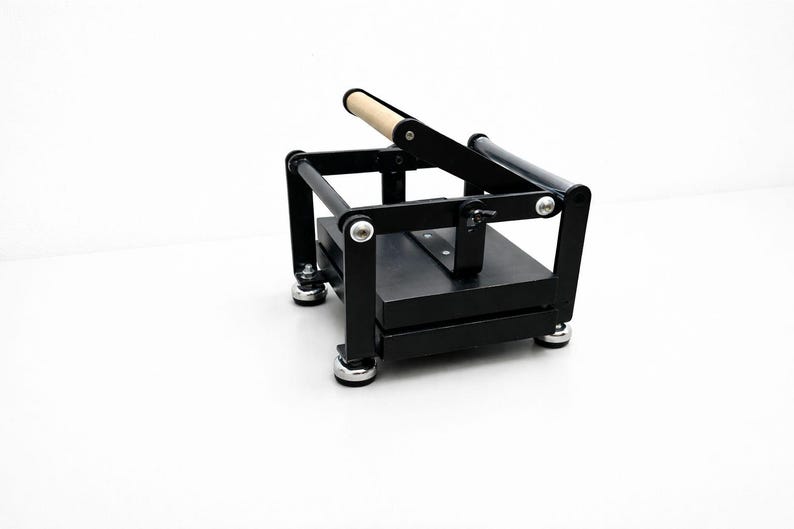 May include: A black book press with a wooden handle. The press has a rectangular base and a hinged top, with silver-colored hardware. The press is designed for crafting or bookbinding projects.
