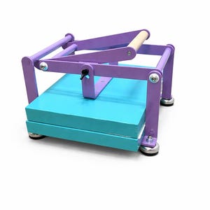May include: A wooden book press painted in purple and teal. The press has a wooden handle and silver hardware. The base is teal and holds a stack of paper. The press is designed for bookbinding and paper crafts.
