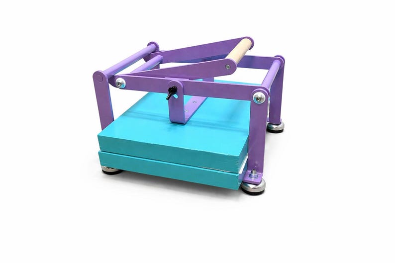 May include: A wooden book press painted in purple and teal. The press has a wooden handle and silver hardware. The base is teal and holds a stack of paper. The press is designed for bookbinding and paper crafts.