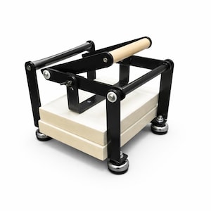 May include: A black and natural wood book press with a light-colored wooden handle. The press has a rectangular base with a stack of light-colored wood blocks. The frame is black metal, and the press sits on four silver feet.