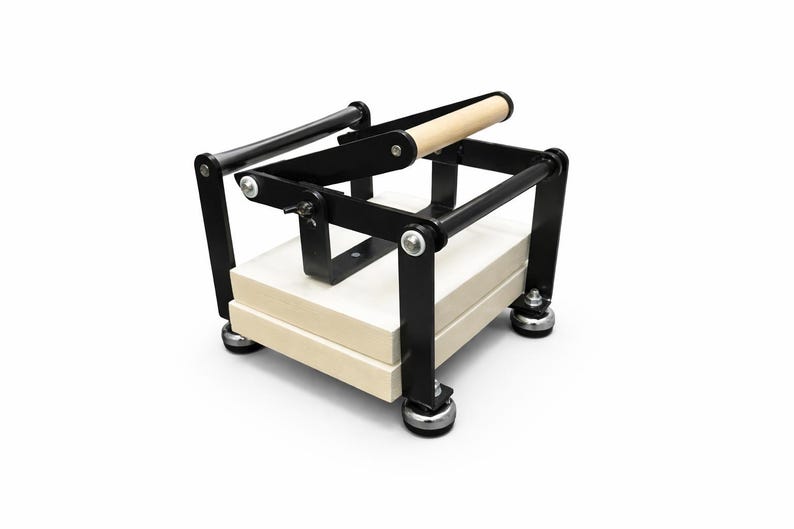 May include: A black and natural wood book press with a light-colored wooden handle. The press has a rectangular base with a stack of light-colored wood blocks. The frame is black metal, and the press sits on four silver feet.