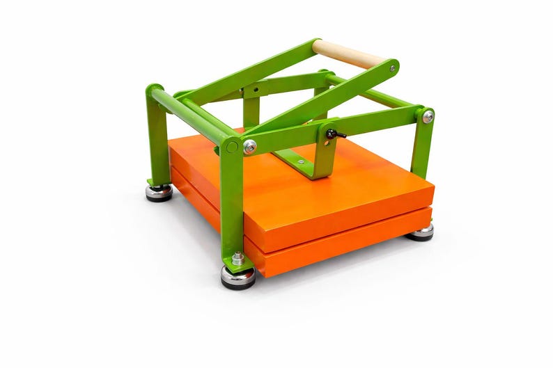 May include: A bright green and orange tortilla press with a wooden handle. The press has a lever design and sits on four small, round feet. The base is a stack of orange wooden boards.