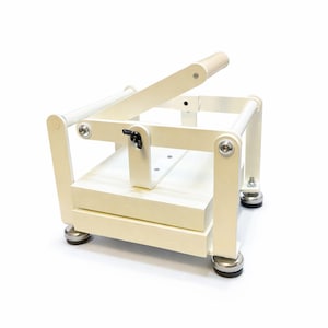 Puede incluir: Un cortador de jabón de madera color crema con una palanca y una cuchilla. El cortador de jabón tiene una base rectangular y un mango. El cortador está diseñado para cortar jabón en barras. El cortador está sobre un fondo blanco.