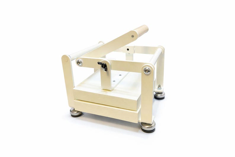 May include: A cream-colored, wooden soap cutter with a lever and a cutting blade. The soap cutter has a rectangular base and a handle. The cutter is designed for cutting soap into bars. The cutter is on a white background.