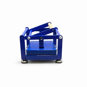 May include: A blue metal press with a wooden handle. The press has a rectangular base and a hinged top. The frame is a vibrant blue, and the handle is a light wood color. The press is designed for crafting or other similar uses.