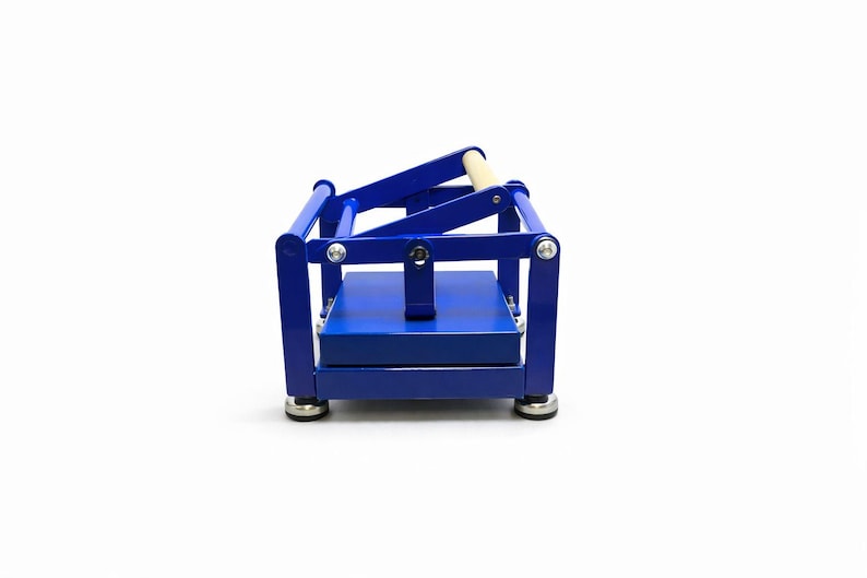 May include: A blue metal press with a wooden handle. The press has a rectangular base and a hinged top. The frame is a vibrant blue, and the handle is a light wood color. The press is designed for crafting or other similar uses.