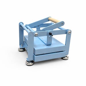May include: A light blue wooden book press with a natural wood handle. The press has a rectangular base and top, connected by vertical supports. The base has four silver-colored feet.
