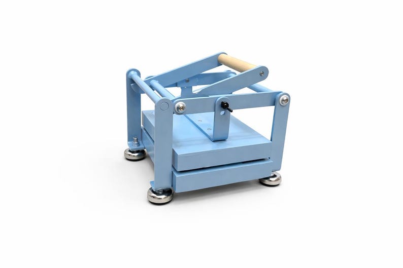 May include: A light blue wooden book press with a natural wood handle. The press has a rectangular base and top, connected by vertical supports. The base has four silver-colored feet.