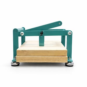May include: A teal-colored book press with a wooden base. The press has a lever arm and a rectangular frame. The base is made of two wooden planks. The press is designed for crafting and bookbinding.