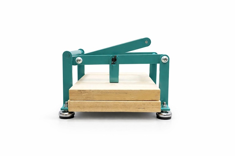 May include: A teal-colored book press with a wooden base. The press has a lever arm and a rectangular frame. The base is made of two wooden planks. The press is designed for crafting and bookbinding.