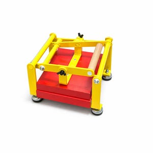 May include: A bright yellow and red printing press with a wooden roller. The press has a rectangular red base and a yellow frame. The press is designed for printmaking and crafting.