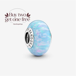 May include: A round Pandora charm bead featuring a smooth, iridescent light blue surface with subtle pink and white flecks. The silver-toned metal core is engraved with the word PANDORA. The text Buy two get one free Harahapshop is visible.