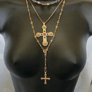 Pode incluir: Conjunto de colares em camadas douradas. A camada superior apresenta uma corrente com pedras preciosas coloridas. A camada do meio tem uma corrente simples. A camada inferior tem um pingente de cruz com pedras preciosas coloridas e um pingente de cruz menor.