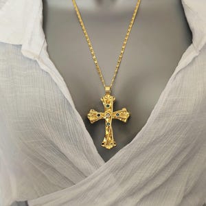 Peut inclure: Un pendentif croix en métal doré suspendu à une chaîne assortie. La croix présente une surface texturée complexe avec de petites pierres circulaires incrustées dans des tons de vert et de bleu au centre et le long des quatre branches.