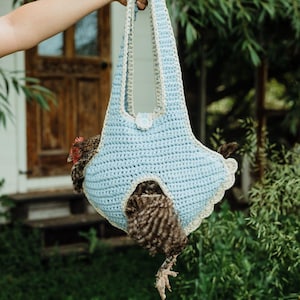 May include: A light blue and cream crochet chicken sling carrier featuring a structured body with a single handle, a scalloped edge trim, and a small button closure. The carrier is designed to hold a brown and black speckled chicken securely.