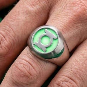 Può includere: Un anello in metallo color argento con una faccia circolare dotata di un inserto luminoso verde chiaro. Il centro dell'anello mostra un simbolo Green Lantern color argento in rilievo, composto da un bordo circolare con una linea centrale.