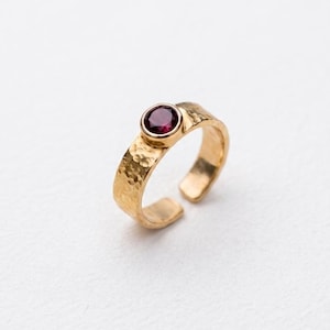 Könnte beinhalten: Ein Goldring mit einem gehämmerten, strukturierten Band und einem einzelnen, runden, tiefroten Granat-Edelstein in einer glatten Fassung. Der Ring hat ein offenes Design zur Anpassung und eine polierte Oberfläche auf dem Metall.