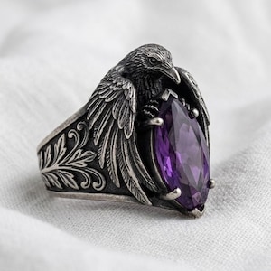 May include: A silver-toned ring featuring a detailed raven sculpture with wings wrapped around a central marquise-cut purple gemstone. The band displays intricate embossed leaf-like patterns along the sides, providing a textured finish.