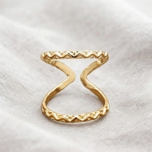 Puede incluir: Un anillo dorado con un diseño de doble banda conectado por una barra vertical central. Tanto la banda superior como la inferior presentan un patrón repetitivo de zigzag o chevrón en los bordes, creando una textura en la superficie.