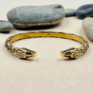 May include: A gold-toned metal cuff bracelet featuring an open design with two symmetrical animal head terminals. The band is decorated with intricate, raised linear patterns and textured detailing along the entire length of the metal surface.