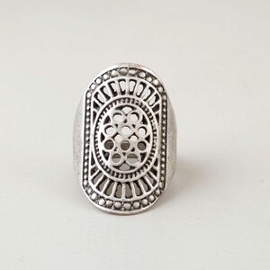 Könnte beinhalten: Ein ovaler, silberfarbener Ring mit einem komplizierten durchbrochenen Design. Die Mitte zeigt ein kreisförmiges Muster aus ineinandergreifenden Schleifen, umgeben von einer Umrandung aus strahlenden Linien und einem Perlenrand, was eine detaillierte Metallstruktur erzeugt.