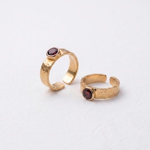Puede incluir: Dos anillos de tono dorado con una textura martillada en la banda y una piedra preciosa granate redonda de color rojo intenso engastada en un bisel. Los anillos son abiertos, lo que permite ajustar el tamaño, y tienen un acabado pulido.