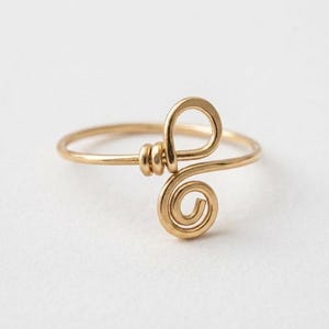 Könnte beinhalten: Ein goldfarbener Drahtring mit einem durchgehenden Band, das eine dekorative Schlaufe und ein Spiralmuster in der Mitte bildet. Der Metalldraht ist um sich selbst gewickelt, um die Form zu sichern, was eine polierte Metalloberfläche am Ring erzeugt.