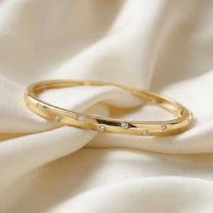 Puede incluir: Una pulsera rígida articulada de tono dorado con acabado pulido y una serie de pequeñas piedras redondas y transparentes incrustadas a intervalos regulares a lo largo de la superficie exterior, creando un efecto brillante sutil y uniforme.