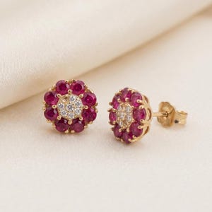 May include: A pair of gold stud earrings featuring a central cluster of small clear stones surrounded by a halo of round, deep red rubies. The floral-shaped design is set in yellow gold, with one earring showing the post and butterfly back.