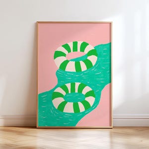 May include: A framed art print featuring two circular pool floats with alternating green and white stripes. The floats are positioned on a textured, wavy turquoise blue surface representing water, set against a solid light pink background.