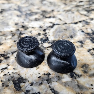 May include: Two black joystick thumbsticks with textured tops. The thumbsticks have a circular design with a raised, ridged pattern. The base of each thumbstick is a smooth, rounded shape.