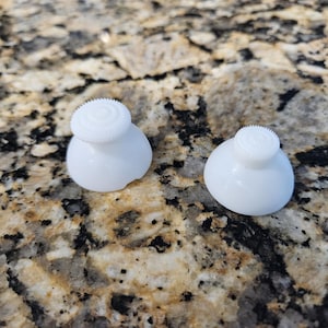 May include: Two white, rounded plastic objects with a textured, circular top. The objects are sitting on a speckled, brown and tan surface. The objects appear to be small knobs or buttons.