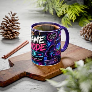 May include: A ceramic coffee mug with a purple handle and rim, featuring a neon-style design with the words "GAME MODE ON" and gaming-related graphics. The mug is filled with coffee and sits on a wooden cutting board.