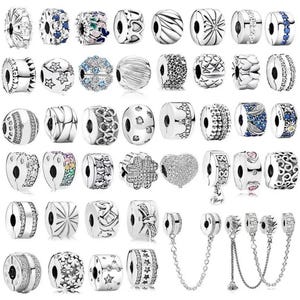Può includere: Una collezione di charm beads argentati con vari design, tra cui motivi floreali, a stella e geometrici. Alcune perline sono adornate con pietre preziose trasparenti o blu, e altre presentano trame intricate. Alcune perline hanno un attacco a catena.