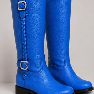 Könnte beinhalten: Ein Paar kniehohe Stiefel in leuchtendem Blau mit strukturierter Oberfläche, einem vertikalen geflochtenen Riemen an der Seite und zwei silberfarbenen Metallschnallen. Die Stiefel haben eine abgerundete Zehenpartie, einen flachen schwarzen Absatz und eine Zuglasche.