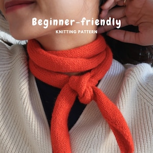 May include: A bright orange knitted scarf tied in a simple knot around the neck, worn over a white ribbed cardigan. The image features the text "Beginner-friendly KNITTING PATTERN" in white letters centred over the orange scarf.