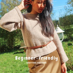 Peut inclure: Un pull en tricot beige à manches longues avec une encolure ronde et des manches évasées, porté avec une fine ceinture en cuir marron dotée d'une boucle dorée. L'image contient le texte blanc "Beginner-friendly KNITTING PATTERN" sur le pull.