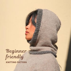 May include: A beige knitted hood with a dark grey ribbed trim around the face opening and neck. The text "Beginner friendly KNITTING PATTERN" is printed in brown on the left side. The hood is made of a soft, textured yarn material.