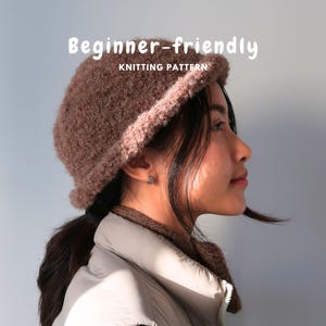 Puede incluir: Un gorro de punto texturizado de color marrón con un borde enrollado. El texto "Beginner-friendly KNITTING PATTERN" aparece en letras blancas sobre el gorro. El gorro presenta una apariencia difusa similar a la lana de color marrón.