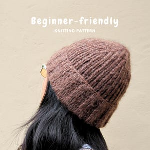 Puede incluir: Un gorro de punto marrón con textura acanalada y borde doblado. El texto "Beginner-friendly KNITTING PATTERN" está impreso en blanco sobre el gorro. El gorro está hecho de hilo grueso y texturizado con un punto de tejido vertical clásico.