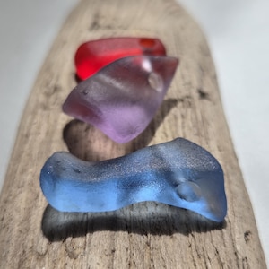 May include: Three pieces of frosted sea glass in red, purple, and blue are arranged in a vertical line. Each piece has a smooth, weathered texture with rounded edges, showcasing translucent colours that catch the light in a soft, diffused manner.