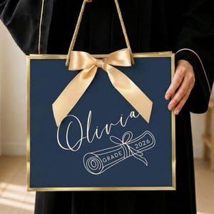 Könnte beinhalten: Eine quadratische marineblaue Geschenktüte mit Goldrand und goldenen Kordelgriffen. Eine große goldene Satinschleife ist oben mittig angebracht. Vorne steht der Name Olivia in weißer Schreibschrift über einer Zeichnung eines Diploms mit GRADE 2026.