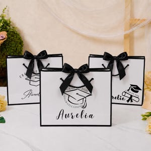 Könnte beinhalten: Drei weiße, rechteckige Geschenktüten mit schwarzem Rand und schwarzen Schleifen. Jede Tüte zeigt eine schwarze Grafik einer Doktorhut-Mütze, den Text "GRAD 2026", "CONGRATULATIONS" sowie den Namen "Aurelia" in schwarzer Schreibschrift.